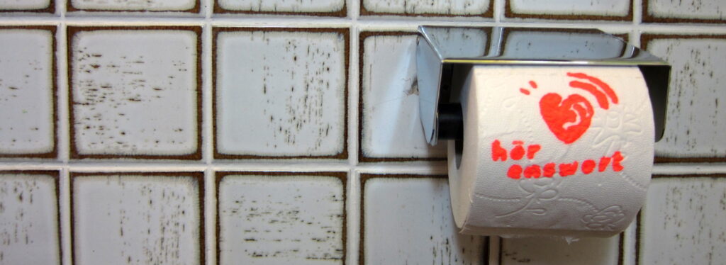 Gekachelte Wand mit einem Toilettenpapierhalter. In diesem ist eine Rolle Klopapier eingespannt, auf die das Logo "HörEnswert" gedruckt ist.
