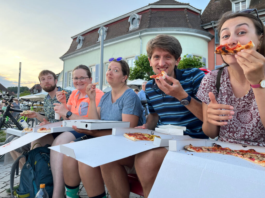 Fünf Personen sitzen auf einer Bank bei Sonnenuntergang und Essen Pizza.